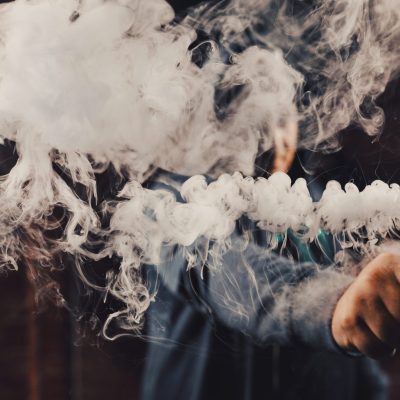 Person holding a smoke-producing wand, thick white vapor swirling around their hands with a blurred figure in the background