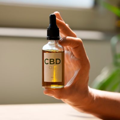 A hand holds a small bottle of CBD oil with a dropper, labeled CBD OIL, backlit by warm light.